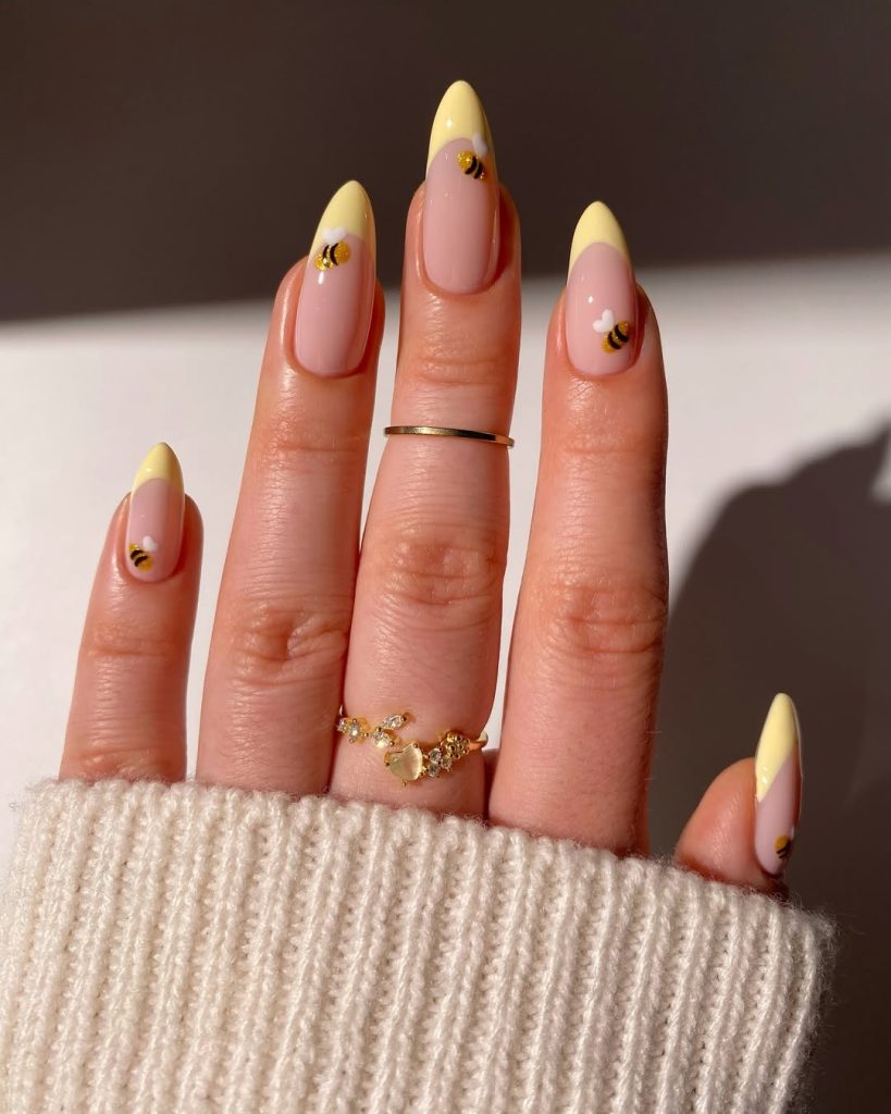 Sheer pink almond nails with yellow French tips and golden bee accents
