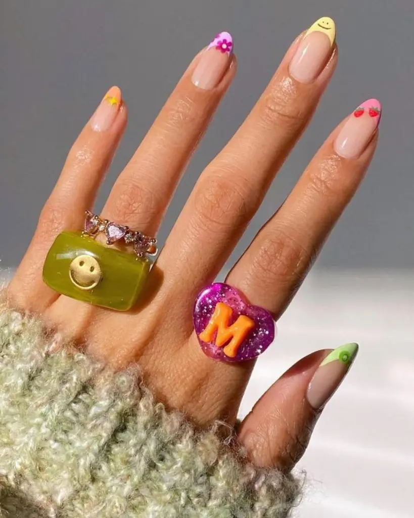 Sheer pink spring nails with colorful French tips featuring smiley faces and flower designs

