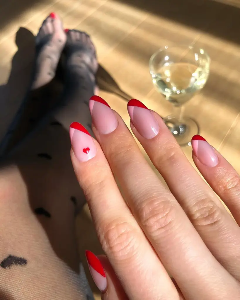 Valentine's Day manicure with red French tips, sheer pink base, and tiny red heart accent nail