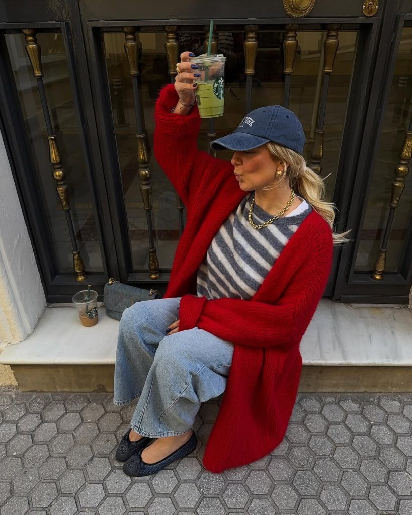 Casual spring outfit featuring a vibrant red oversized cardigan layered over a gray and white striped top, light wash straight-leg jeans, quilted black flats, navy baseball cap, chunky gold chain necklace, and a small gray handbag
