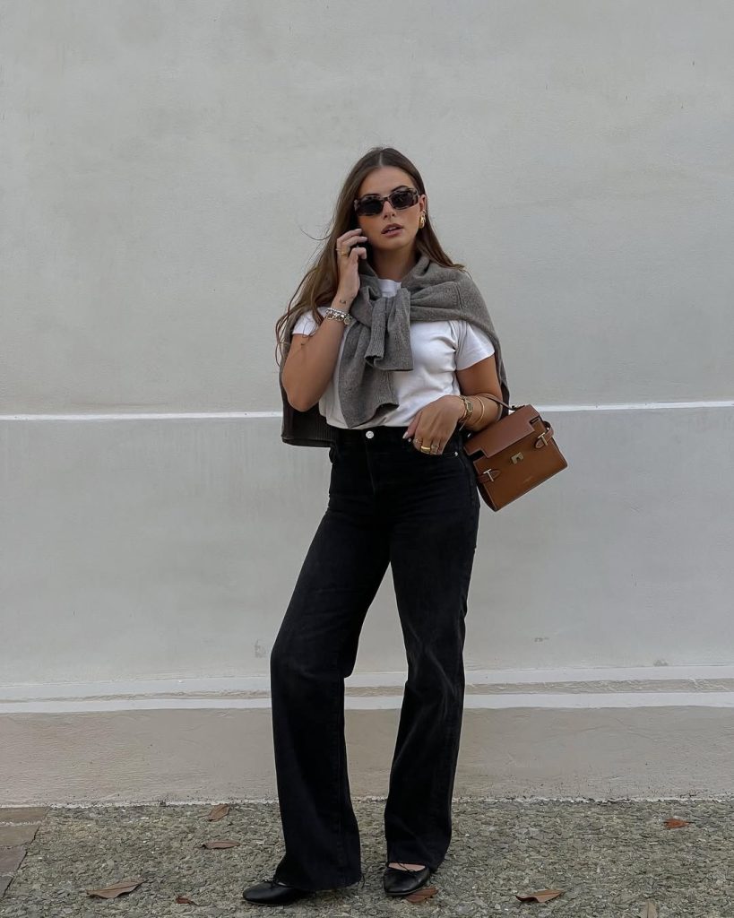 Everyday spring outfit featuring a crisp white t-shirt, black wide-leg trousers, gray cashmere sweater draped over shoulders, structured tan leather handbag, black sunglasses, and gold accessories
