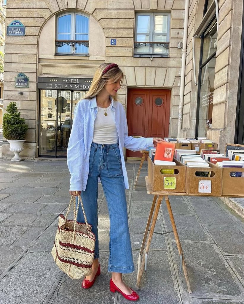 Casual spring outfit featuring a light blue button-down shirt over white fitted tank top, high-waisted wide-leg utility jeans, bright red block heel shoes, woven straw tote bag, and burgundy headband
