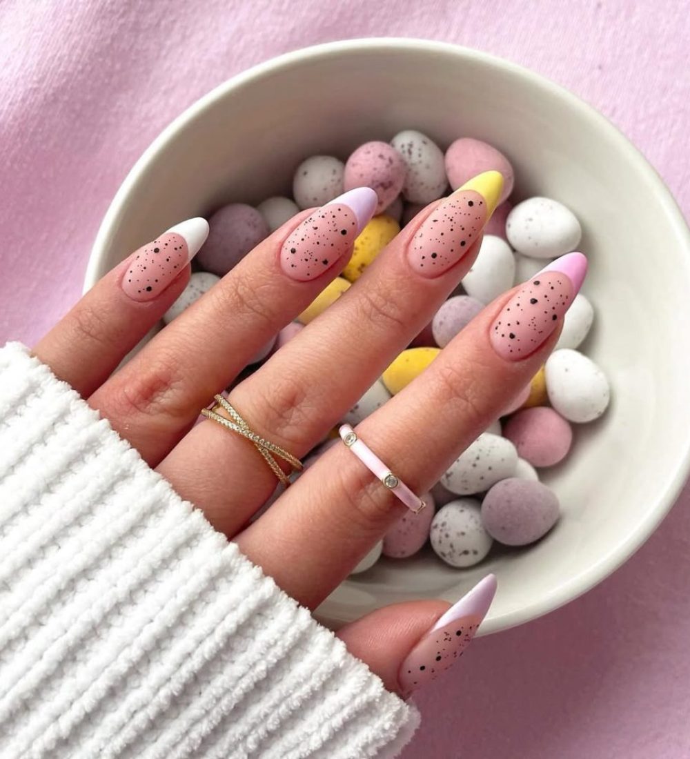 Almond-shaped pink nails with colorful pastel French tips in white, yellow, and lavender, decorated with black speckles throughout for a contemporary Easter egg-inspired manicure