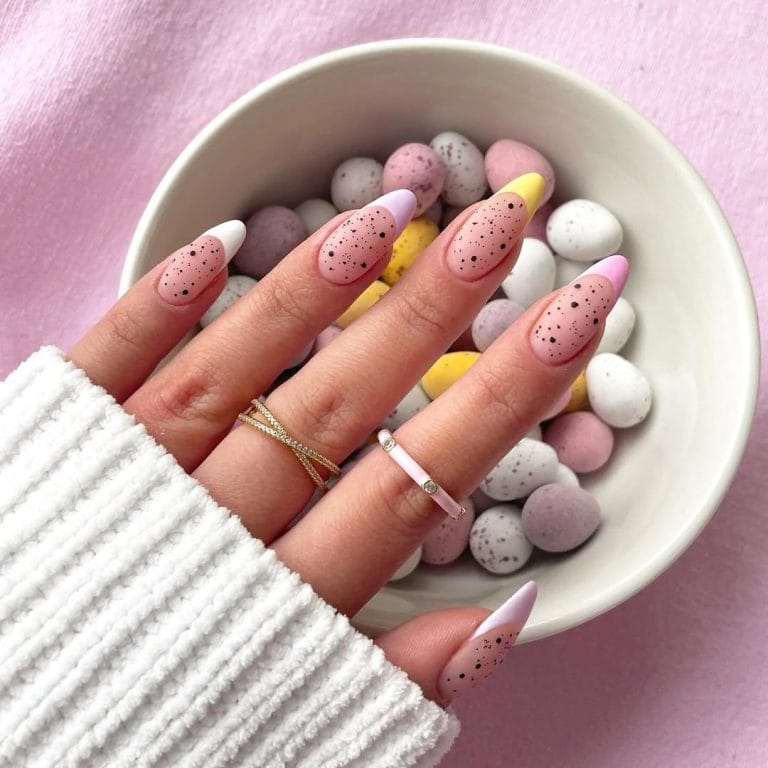 Almond-shaped pink nails with colorful pastel French tips in white, yellow, and lavender, decorated with black speckles throughout for a contemporary Easter egg-inspired manicure