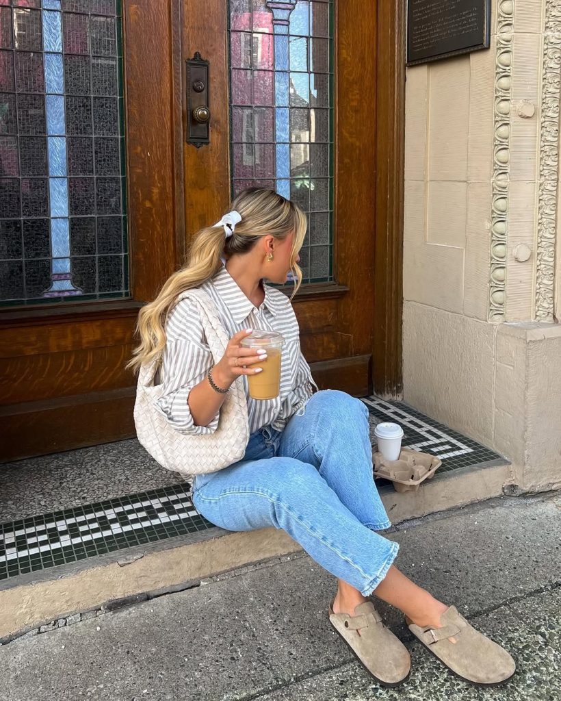 Casual spring outfit featuring a gray and white striped button-down shirt, light wash straight-leg jeans, beige suede clogs, cream woven shoulder bag, and white hair scrunchie
