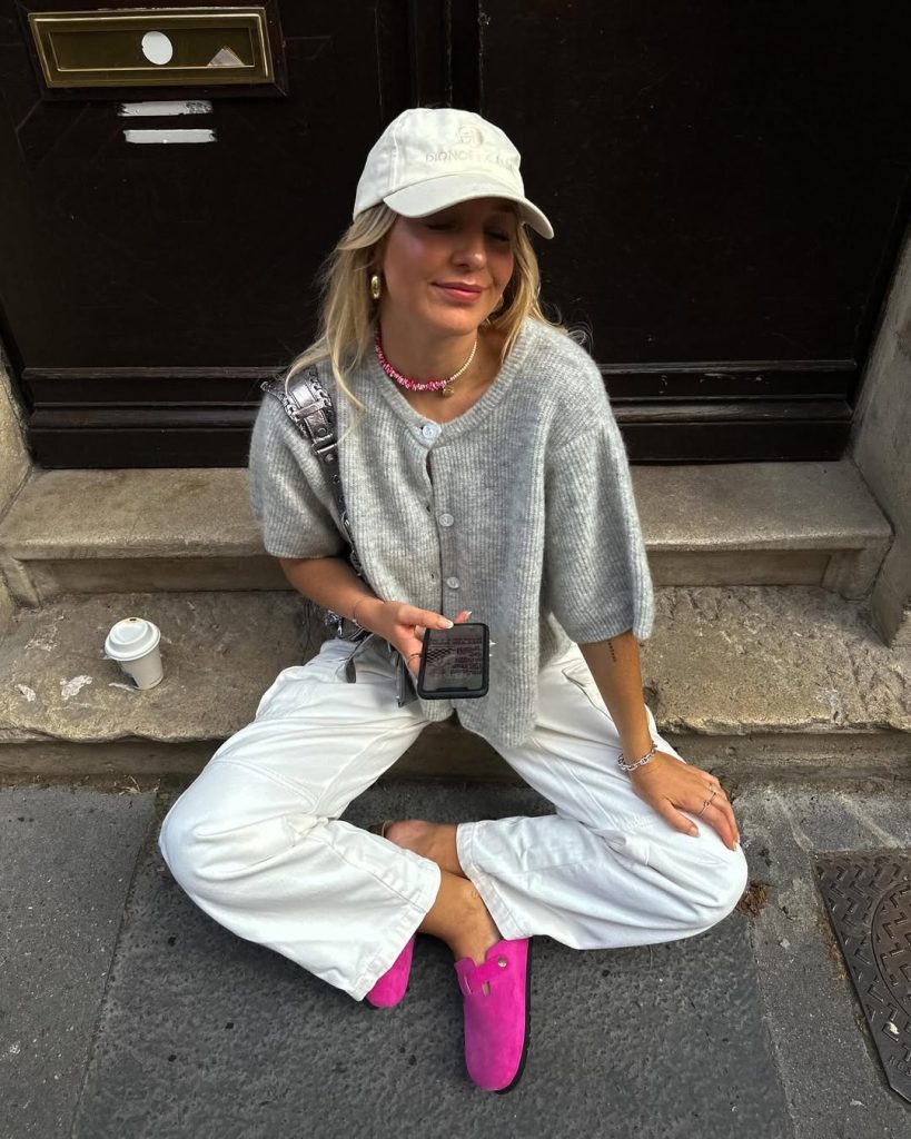 Casual spring outfit featuring a light gray short-sleeve button-up cardigan, white wide-leg jeans, bright pink mules, cream baseball cap, beaded necklace, and silver crossbody bag
