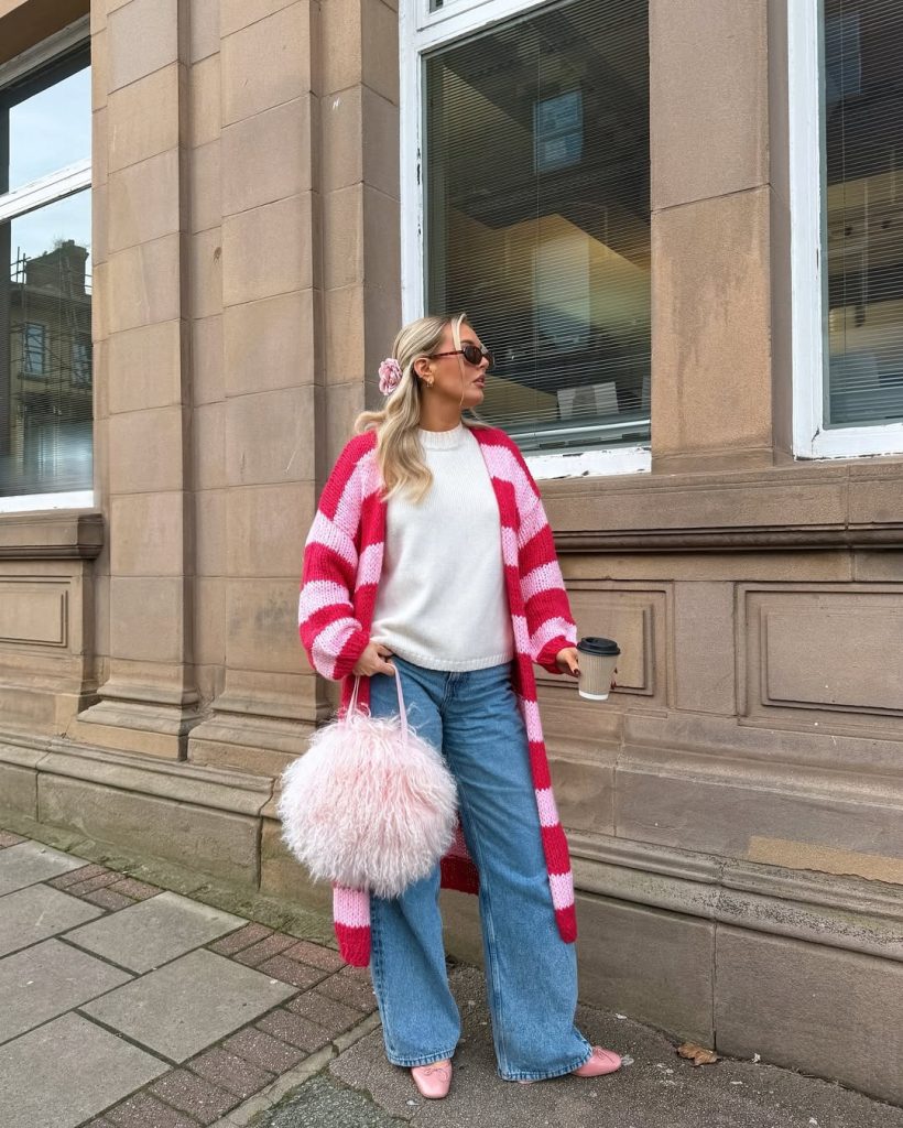 Playful spring outfit featuring a long pink and white striped knit cardigan over cream sweater, relaxed light wash jeans, pink ballet flats, fluffy pink handbag, and coordinating pink scrunchie
