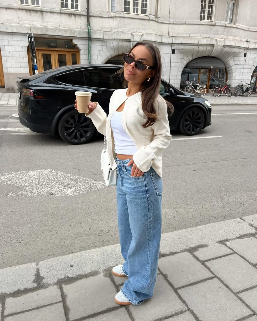 Casual spring outfit featuring a cream cardigan, white fitted crop top, light wash wide-leg jeans, white sneakers with gum soles, small white shoulder bag, and oval sunglasses
