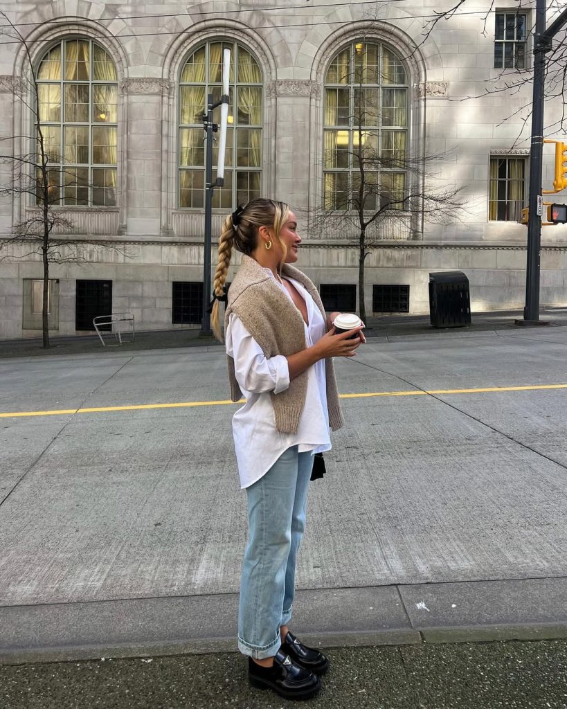 Classic spring outfit featuring a beige knit sweater draped over shoulders, oversized white button-down shirt, light wash straight-leg jeans, chunky black loafers, small black shoulder bag, and pearl necklace for an effortlessly elegant everyday look
