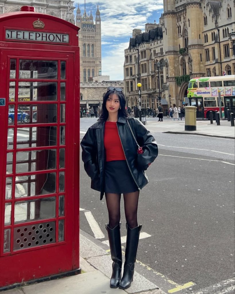 Woman in red top, leather jacket and mini skirt with boots near London phone booth
