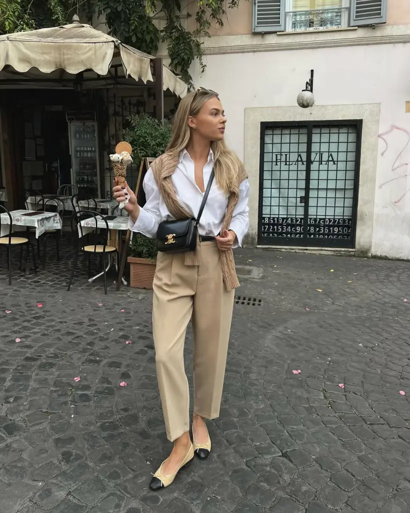 Classy outfit with white button-down shirt, draped camel sweater, beige tailored trousers, black and beige ballet flats and small black crossbody bag