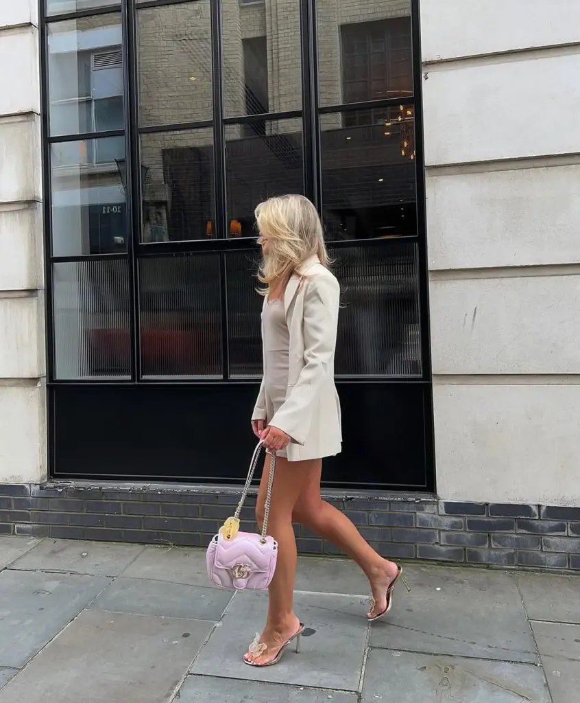 Woman in cream blazer dress with clear heels and pink designer bag on city street