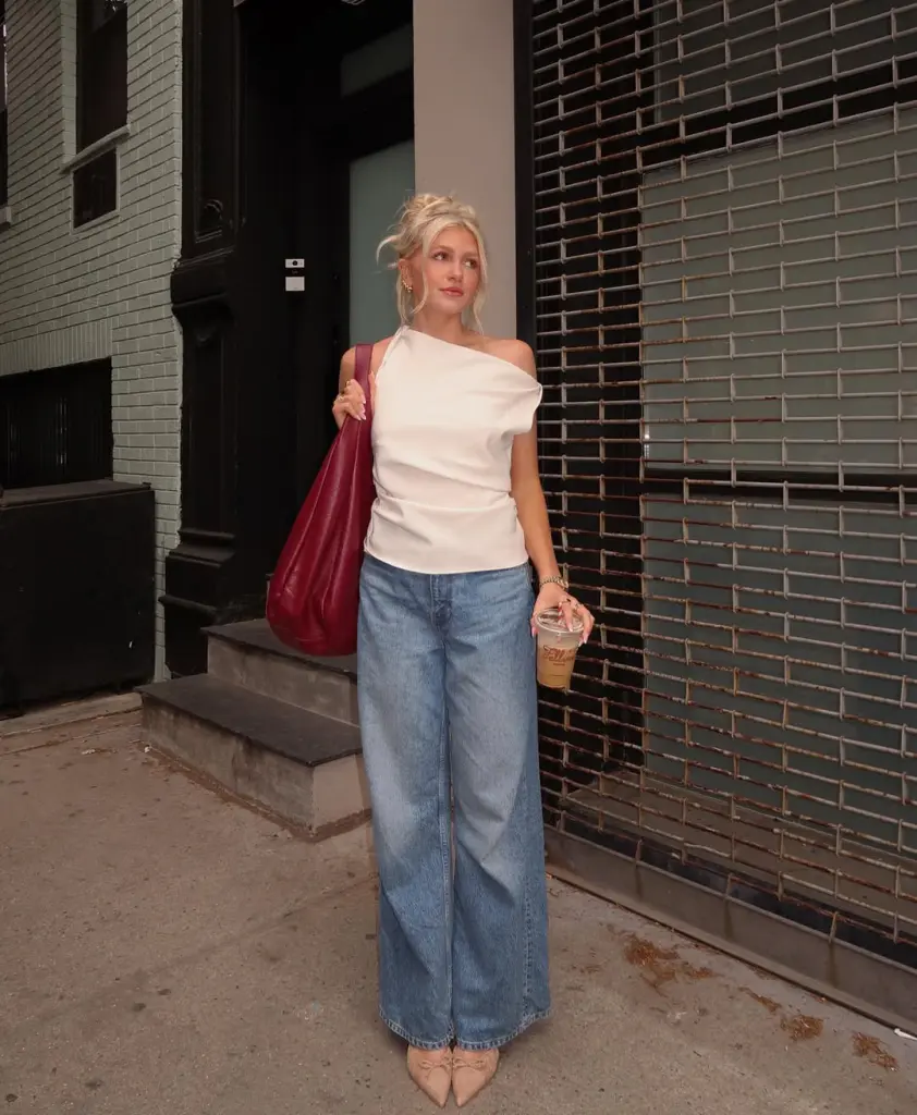 Woman in white draped top with light wash wide leg jeans and nude flats carrying burgundy bag