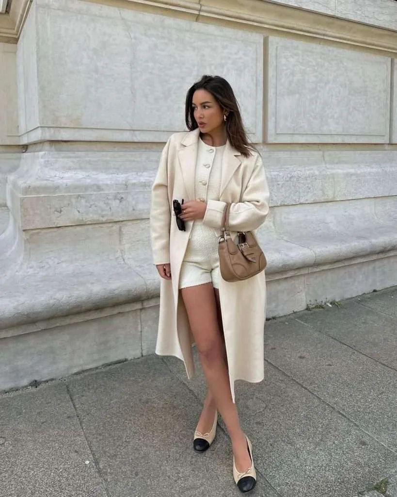 Classic spring outfit with cream long coat, tailored ivory high-waisted shorts, black cap toe ballet flats and beige leather top handle satchel bag