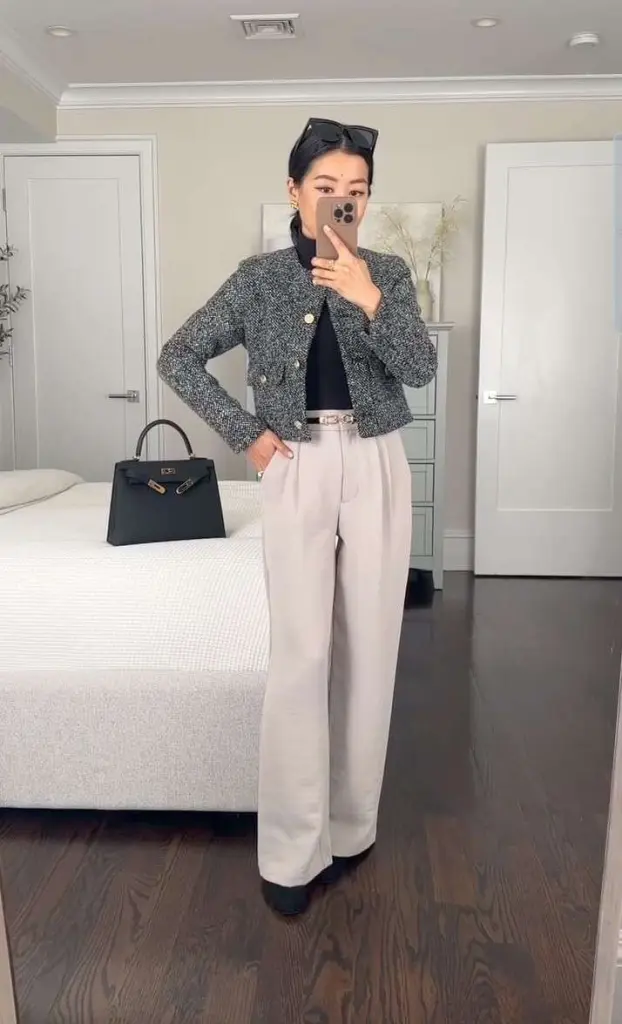 Business casual outfit with grey tweed cropped jacket, black top, cream wide-leg trousers, belt, and structured black handbag in bedroom setting