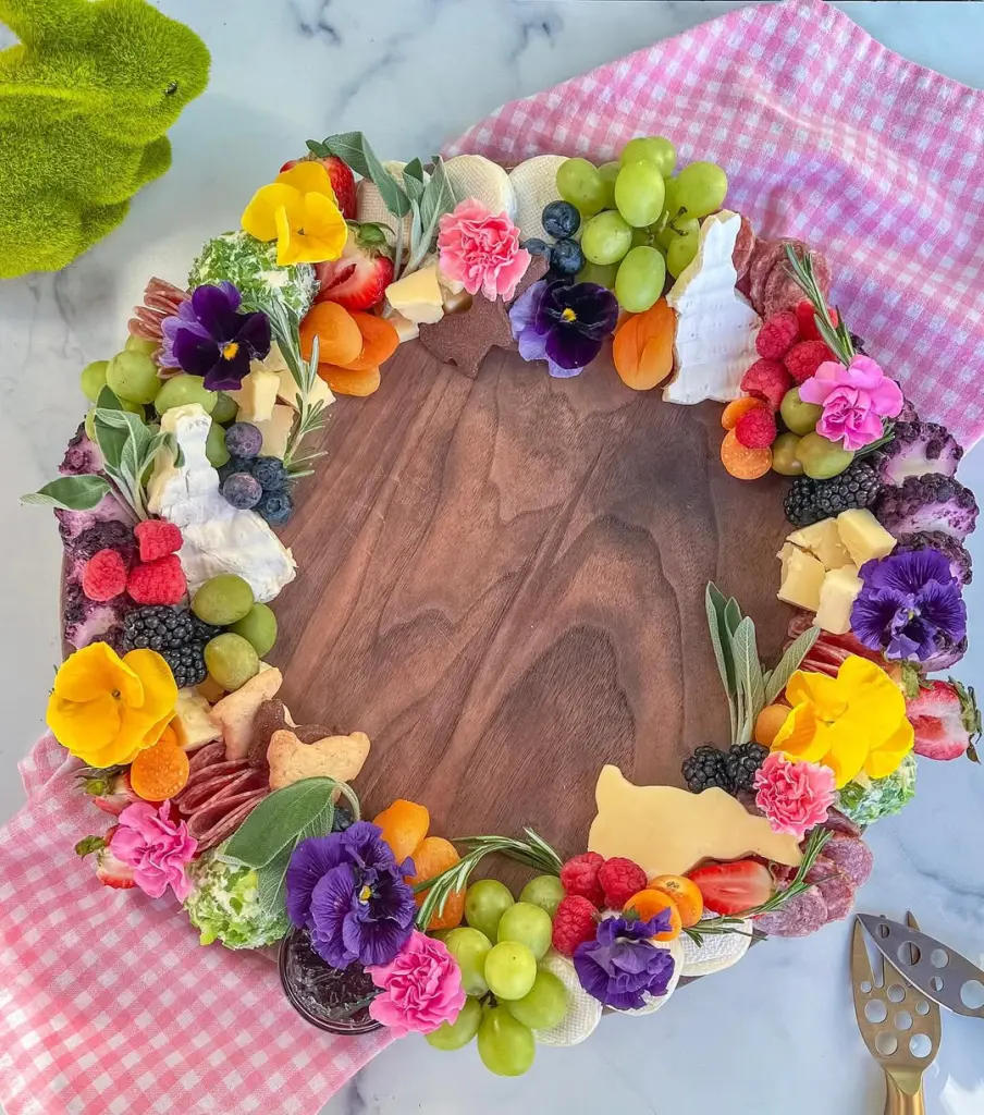 Easter charcuterie wreath featuring seasonal fruits, cheeses, edible flowers, bunny-shaped crackers, and charcuterie arranged in a circular pattern on a wooden board with pink gingham napkin