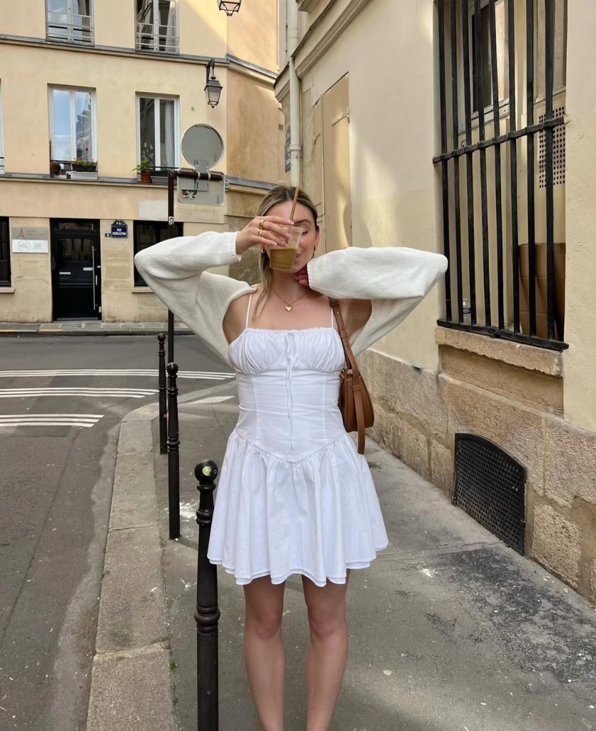 Feminine spring outfit with white ruched sundress, cream knit cardigan, leather crossbody bag, gold pendant necklace