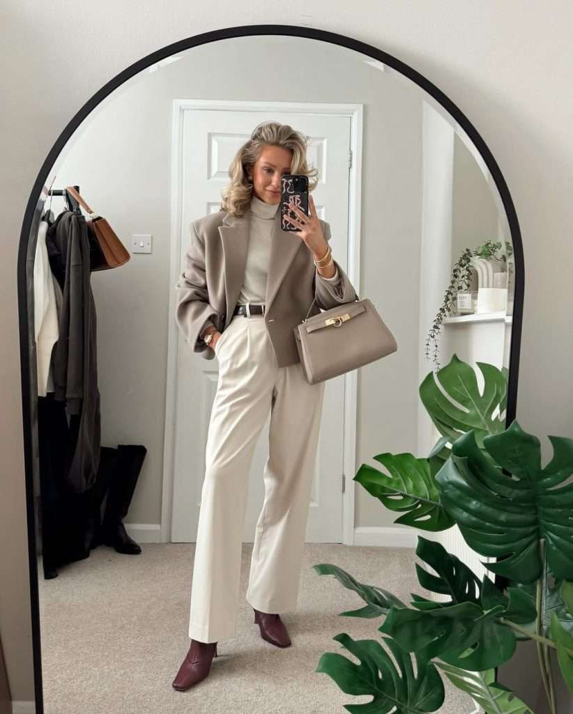 Business casual outfit with oversized taupe blazer, cream turtleneck, white wide-leg trousers, black belt, burgundy boots and beige structured handbag in mirror selfie