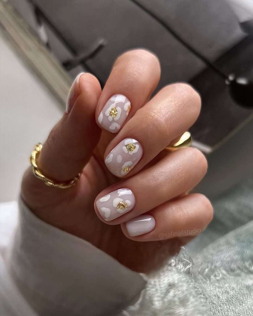 Short square nails with nude base, white flower designs and gold foil accents
