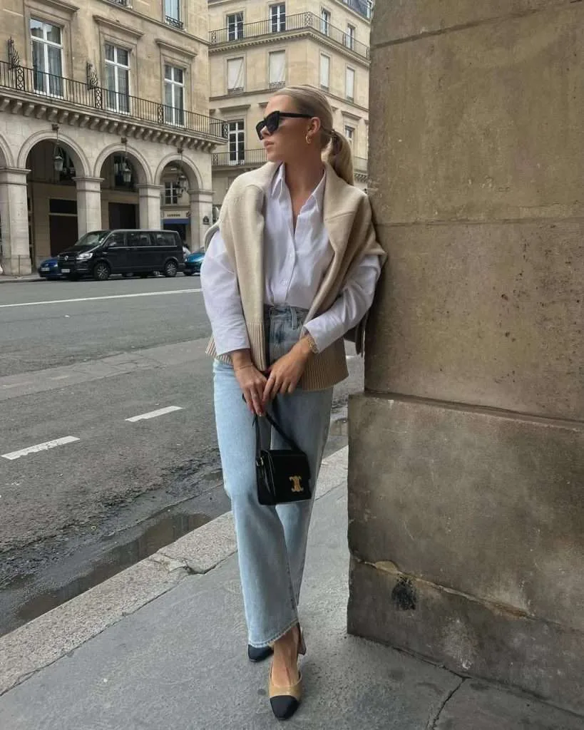 Classy spring outfit with cream draped sweater, white button-down shirt, light wash jeans, two-tone flats and black Celine bag