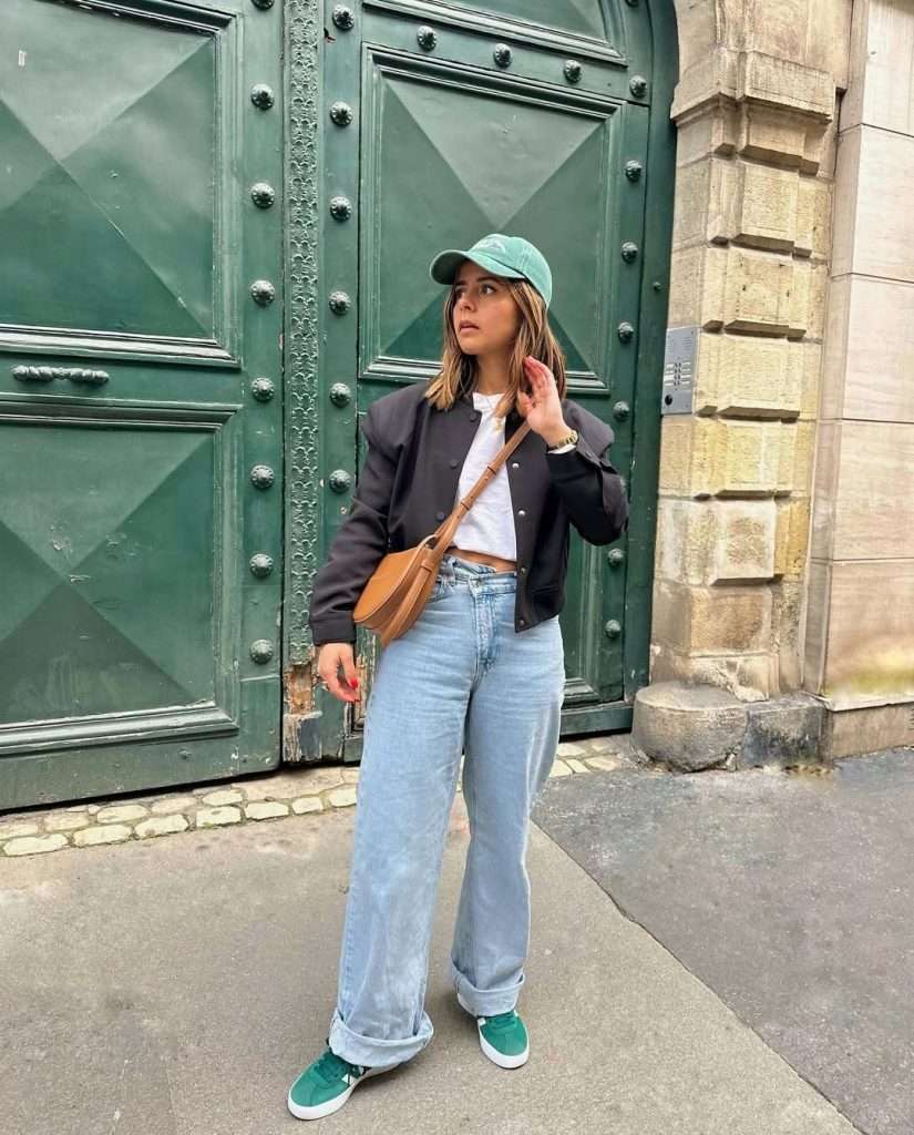 spring outfit with black bomber jacket, white crop top, wide leg jeans, mint green baseball cap, green sneakers and tan crossbody bag
