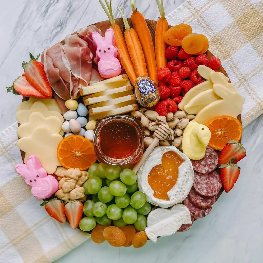 Easter charcuterie board featuring Peeps marshmallow bunnies, fresh carrots, honey, cheese cutouts, raspberries, strawberries, grapes, and chocolate egg surrounded by prosciutto and salami on a wooden platter