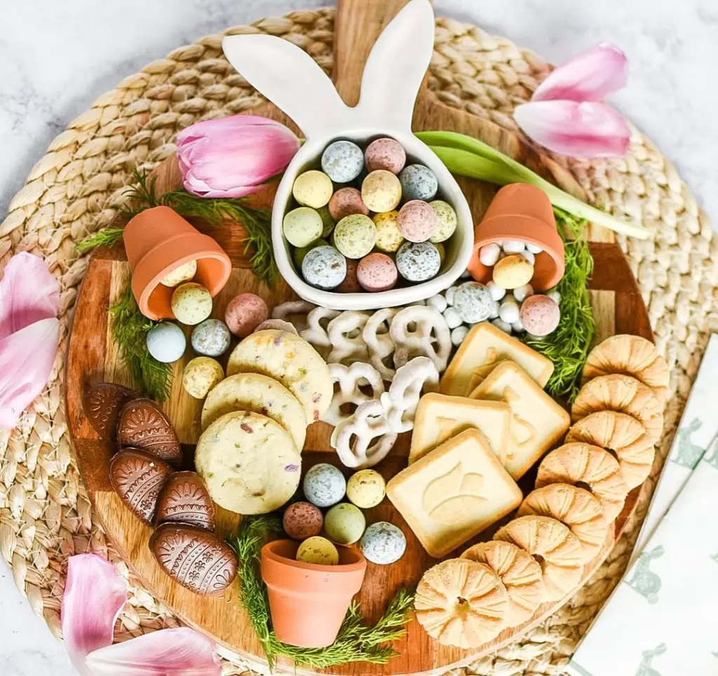 Elegant Easter-themed charcuterie featuring bunny-shaped dish with candy eggs, terracotta pots with treats, chocolate eggs, yogurt-covered pretzels, and spring-themed cookies arranged on a wooden platter with tulip accents