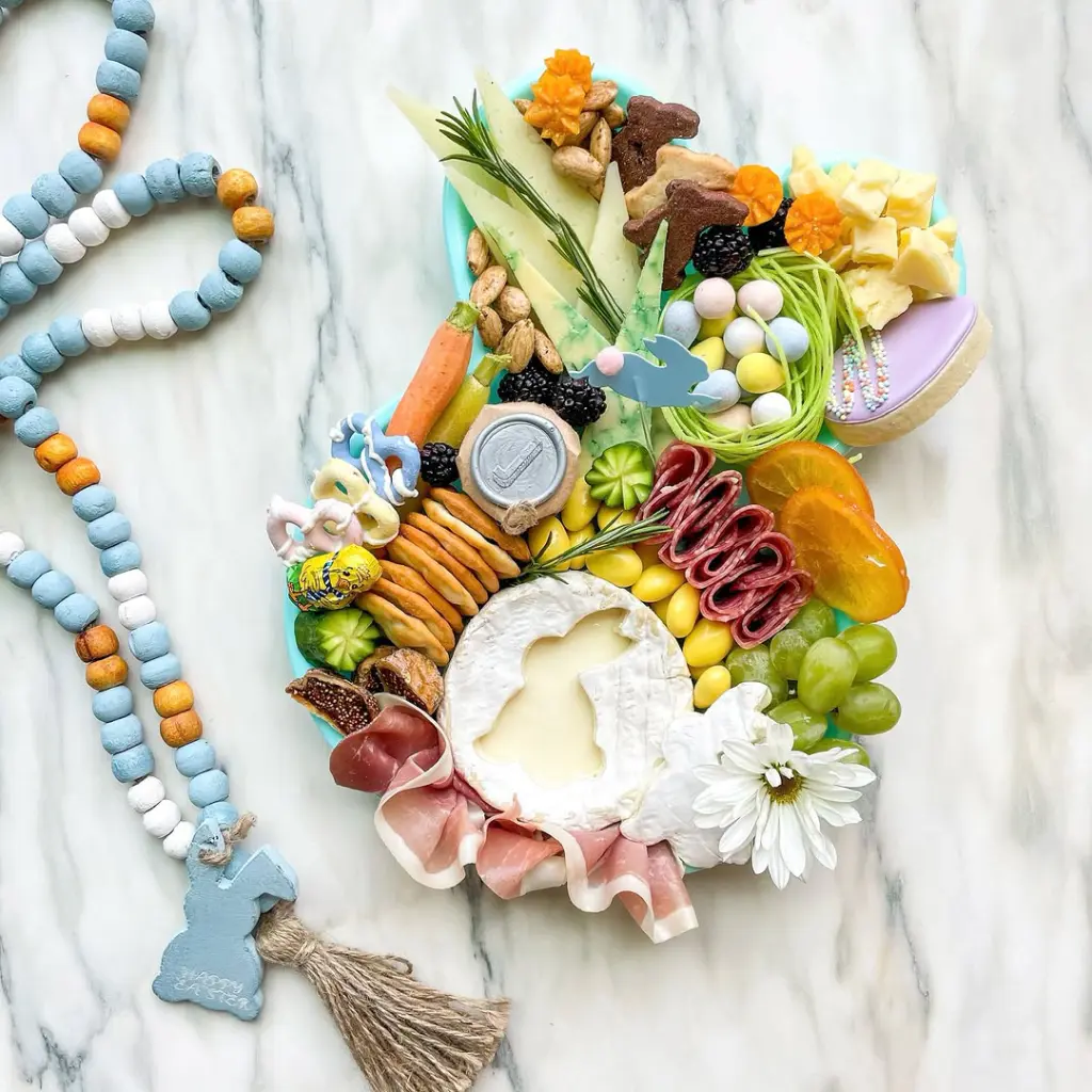 Easter charcuterie board with bunny-shaped brie centerpiece, fresh fruits, crackers, salami, bunny cookies, candy eggs in a grass nest, and decorative wooden bead garland on a blue serving tray