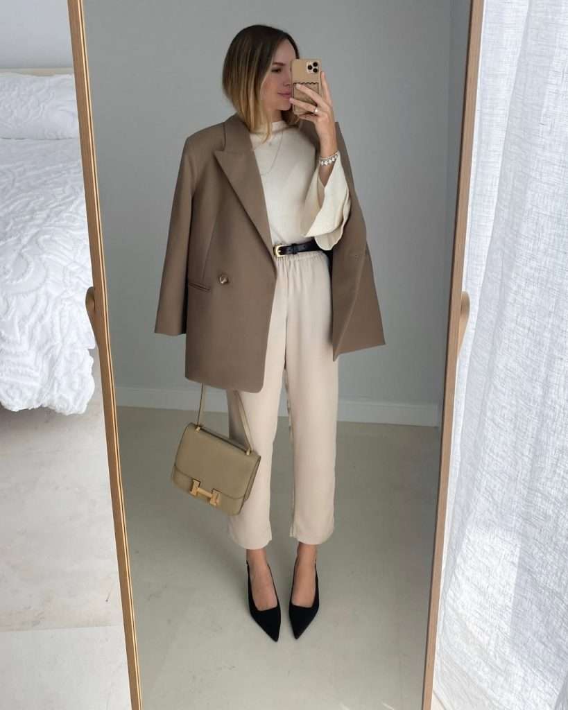 Business casual outfit with camel oversized blazer, cream sweater, cream ankle-length trousers, black belt, black pointed pumps and taupe structured handbag