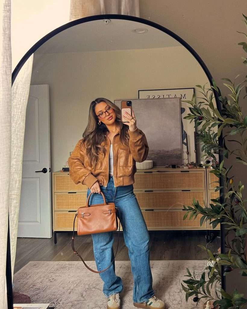  spring outfit with tan leather bomber jacket, white t-shirt, medium wash straight leg jeans, beige sneakers and tan leather handbag