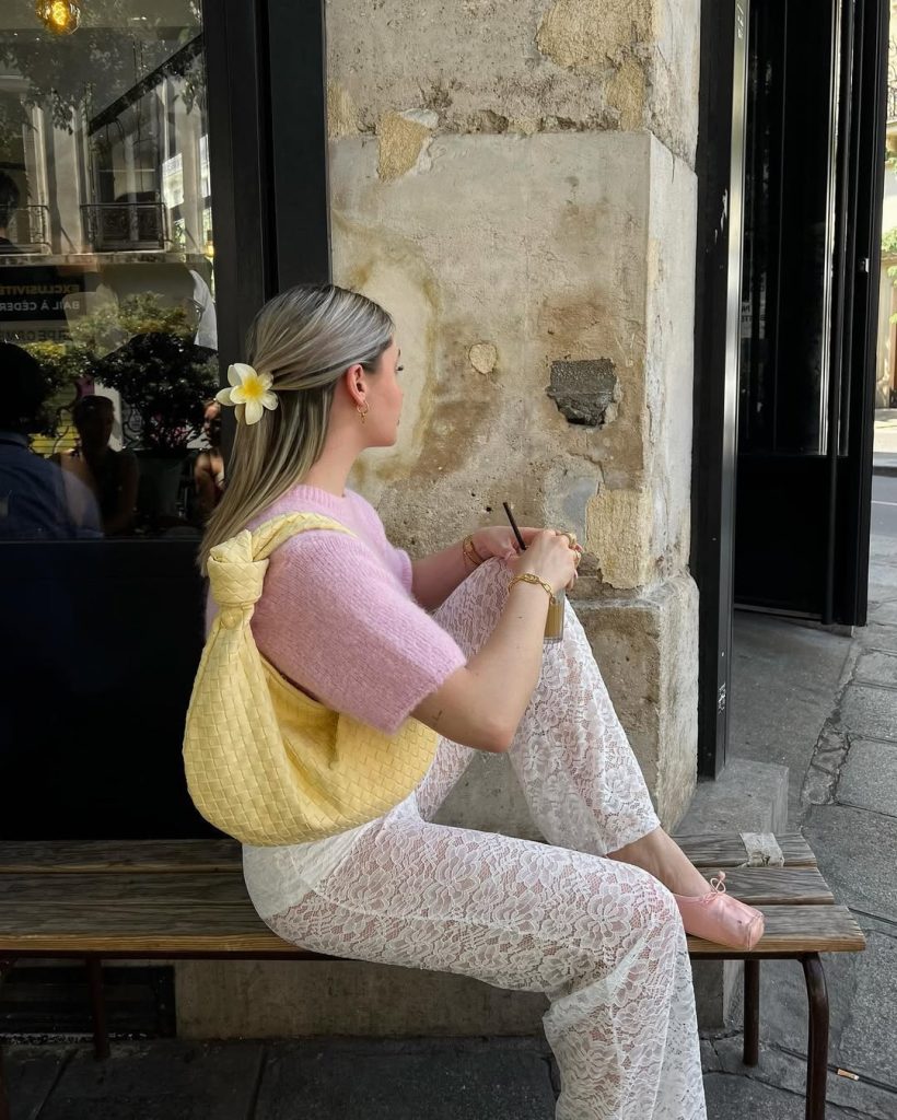 Feminine spring outfit with pink short-sleeve sweater, white lace pants, pink ballet flats, yellow woven tote bag and flower hair accessory