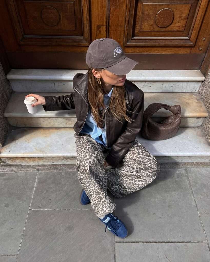 Casual spring outfit with brown leather jacket, light blue t-shirt, beige leopard print wide leg pants, black baseball cap and brown hobo bag
