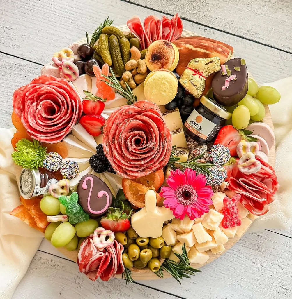 Easter charcuterie board featuring salami roses, chocolate eggs, bunny-shaped cheese and cookies, green olives, strawberries, pink gerbera daisy, and assorted cheeses on a wooden platter with rustic backdrop
