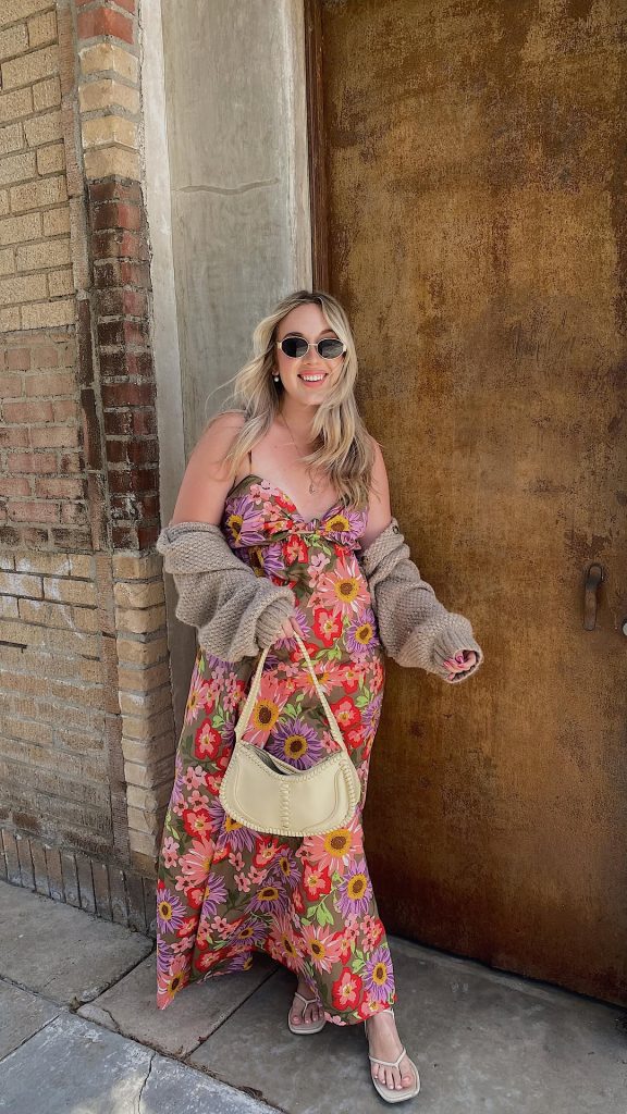 Colorful spring outfit with pink and purple floral print maxi dress, taupe chunky knit cardigan, cream shoulder bag, flip flop sandals and round sunglasses
