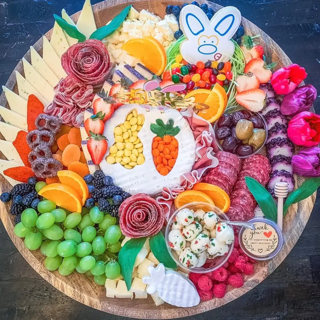 Easter charcuterie board featuring brie with yellow and orange candy arranged as carrot and bunny shapes, decorated sugar cookie, fresh fruits, salami roses, pretzels, cheese slices, and edible flowers on a wooden platter