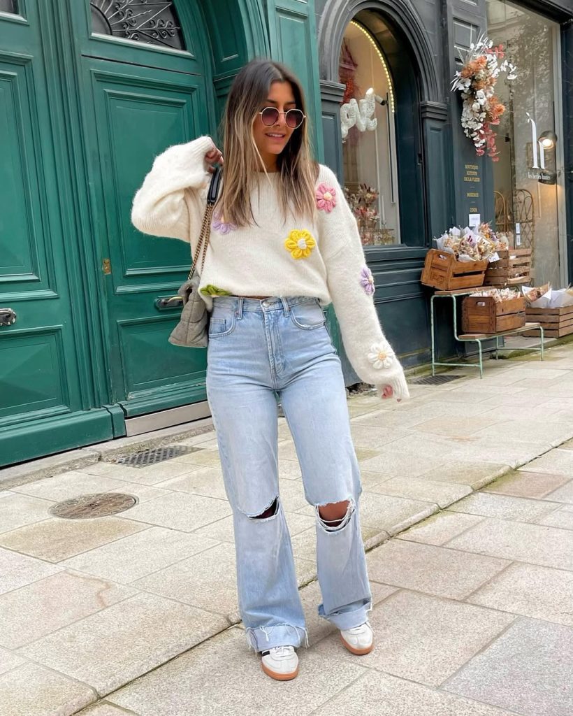 Casual spring outfit with cream knit sweater with colorful flower appliqués, light wash distressed wide-leg jeans, white sneakers, grey shoulder bag and round sunglasses
