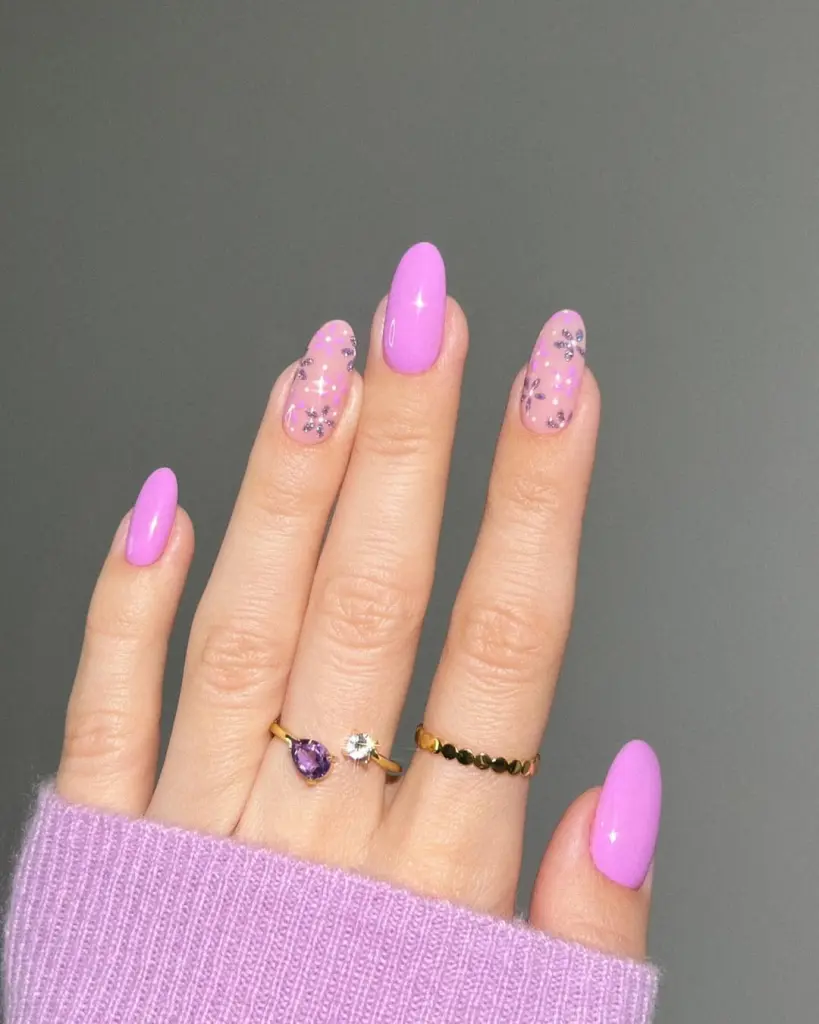  Almond shaped nails with bright lilac polish alternating with transparent pink featuring purple flowers and gold glitter details
