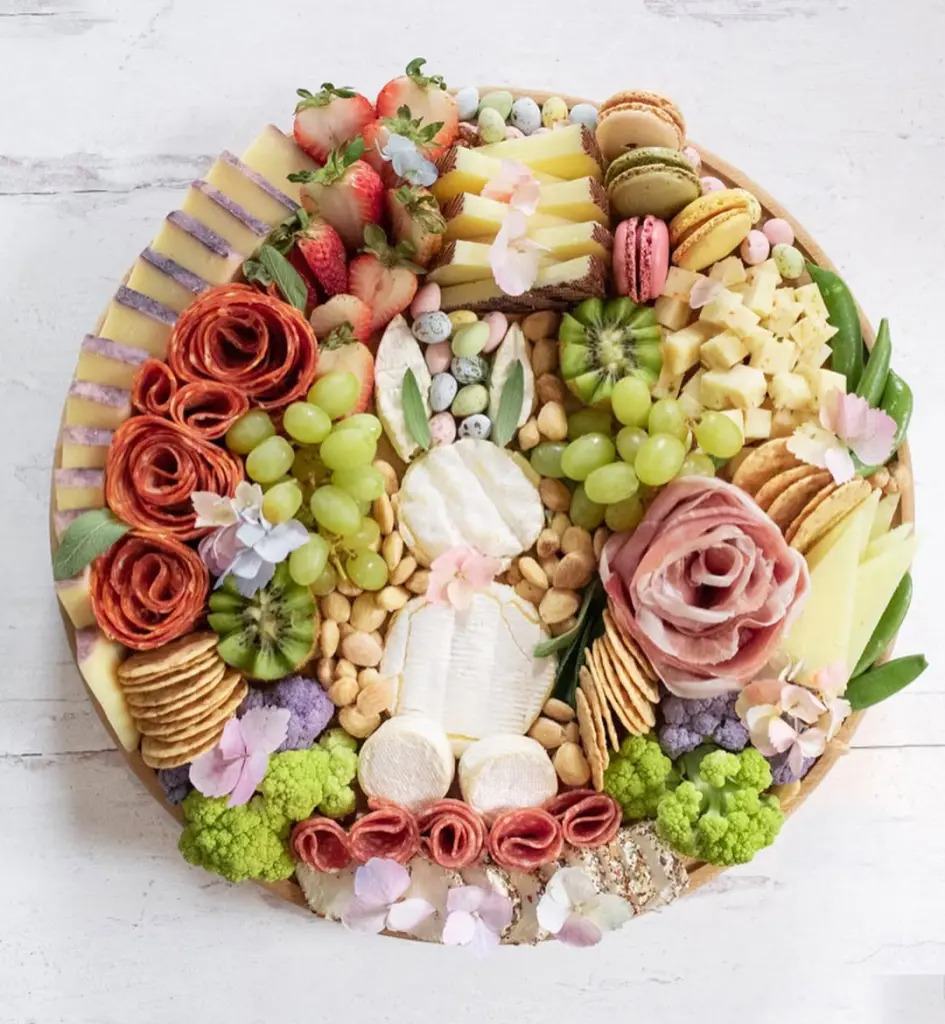 Elegant Easter charcuterie board with soft cheese flowers, prosciutto roses, salami roses, fresh fruits, pastel candy eggs, edible flowers, crackers, and colorful macarons arranged on a wooden circular tray