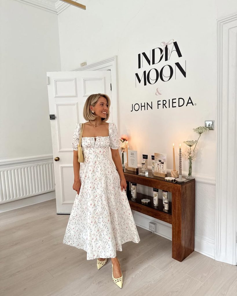 Elegant spring outfit with white floral print midi dress with puff sleeves and square neckline, pale yellow studded pointed flats, gold layered necklaces and small shoulder bag
