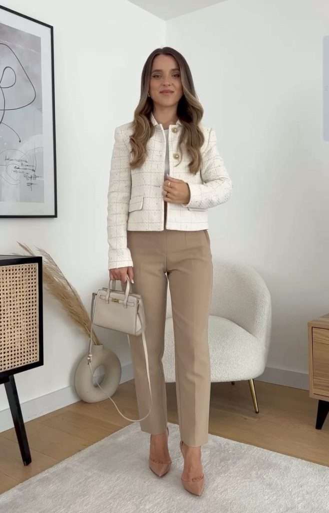 Professional outfit with cream tweed jacket, light pink top, camel straight-leg trousers, nude pumps and matching mini structured handbag in minimalist living room. 
