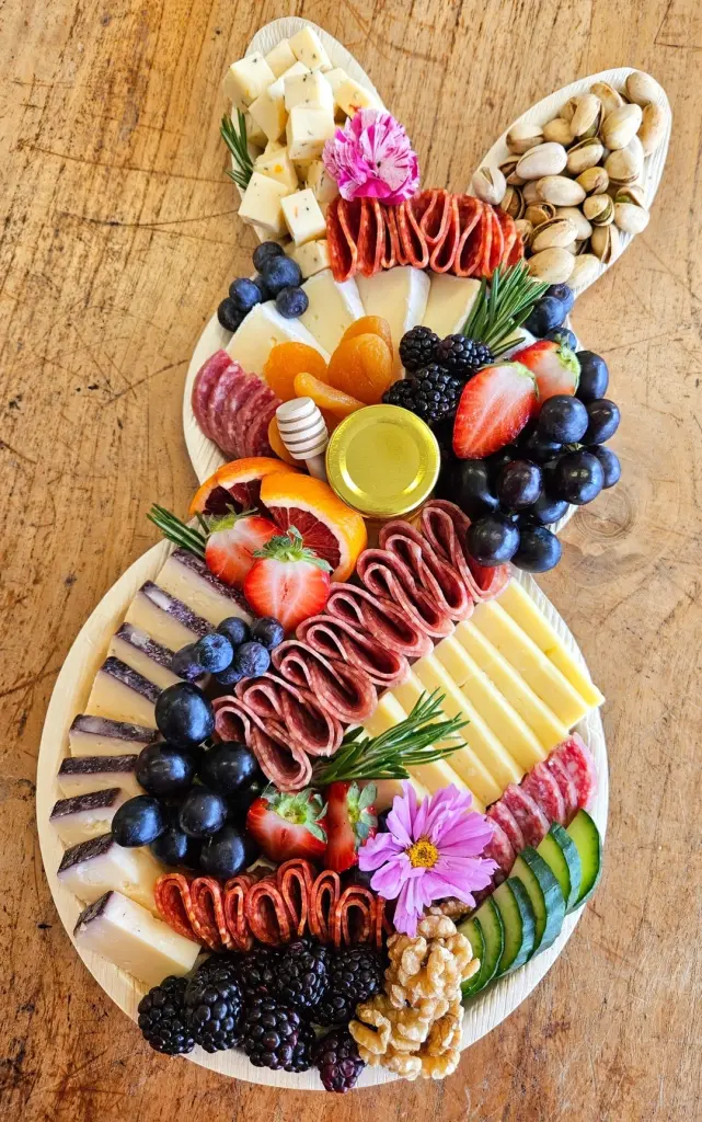 Easter bunny-shaped charcuterie board with sliced cheeses, salami, fresh berries, grapes, pistachios, honey, and edible flowers arranged on a wooden platter with rosemary sprigs
