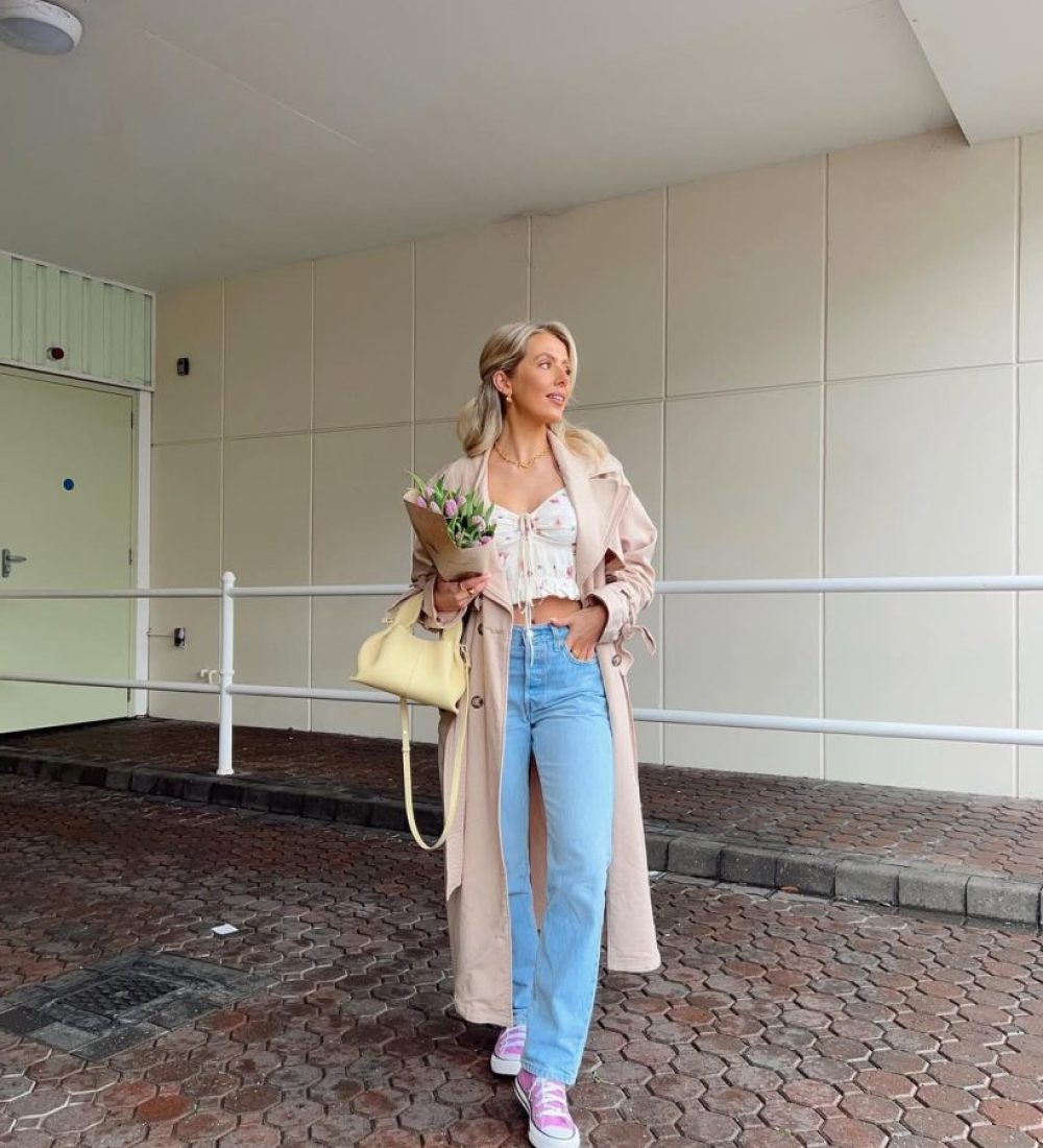 Casual spring outfit with white floral print crop top, light wash straight leg jeans, beige trench coat, pink canvas sneakers and pale yellow shoulder bag