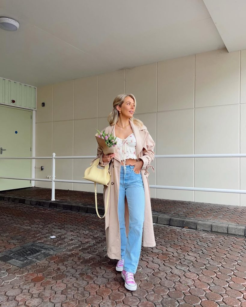 Casual spring outfit with white floral print crop top, light wash straight leg jeans, beige trench coat, pink canvas sneakers and pale yellow shoulder bag
