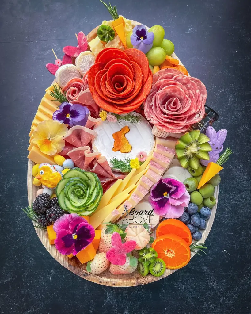 Oval Easter charcuterie board with brie featuring carrot bunny silhouette, salami roses, cucumber rose, fresh fruits, vibrant edible flowers, cheese slices, and colorful peeps arranged on wooden platter