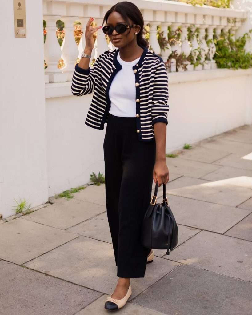 Business casual outfit with navy and white striped cardigan, white top, black straight-leg trousers, beige and black cap-toe flats and black bucket bag

