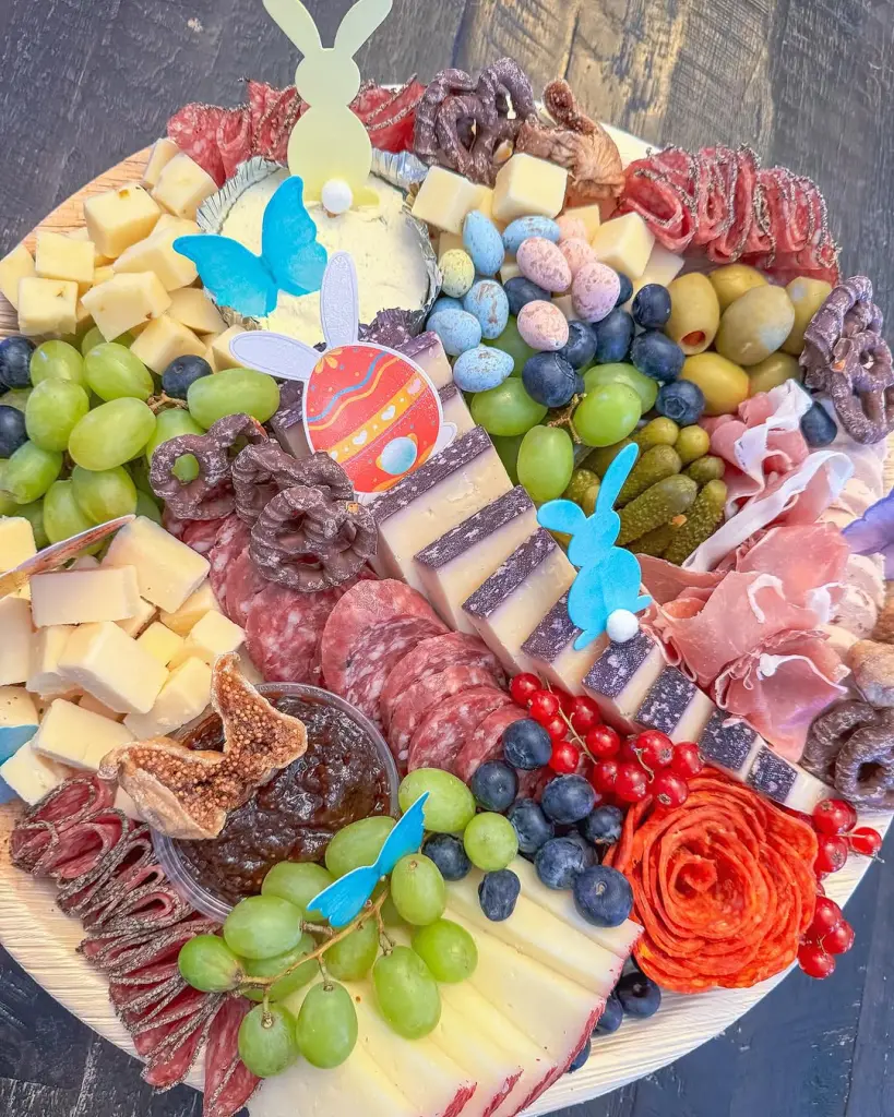 Easter charcuterie board with colorful cheese slices, cured meats, speckled candy eggs, grapes, olives, blueberries, fig jam and decorative blue butterfly and bunny silhouettes on a wooden platter