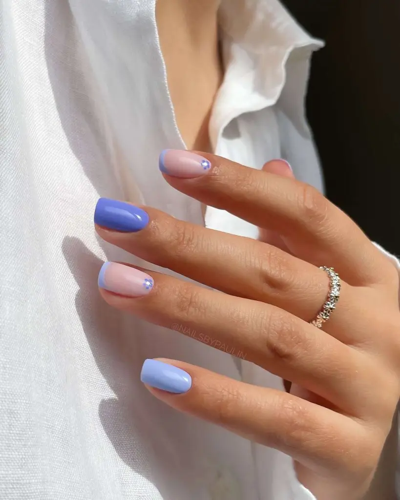 Short oval nails with alternating periwinkle polish and milky base with blue French tips and flower accents
