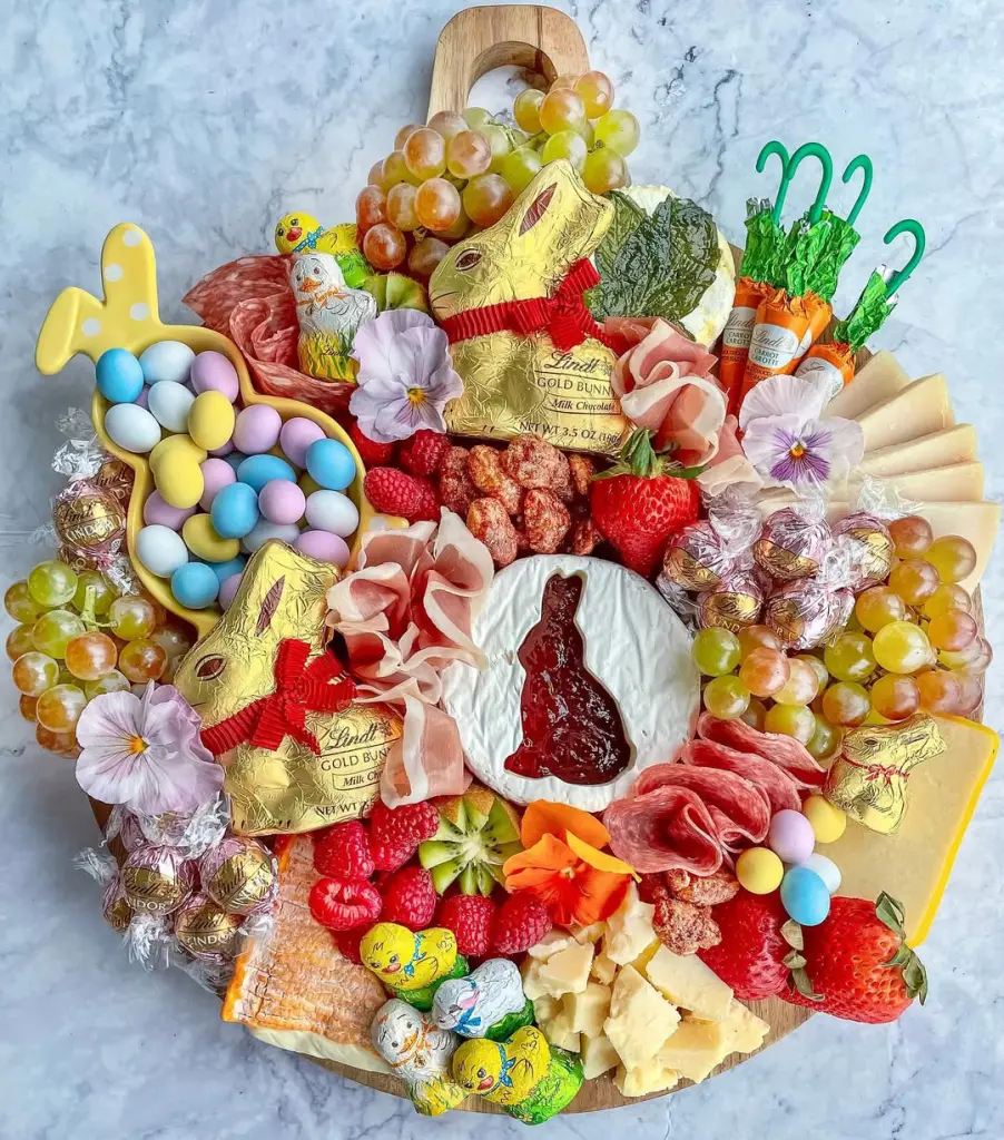 Easter charcuterie board featuring Lindt gold bunnies, bunny-shaped cheese with jam, bunny-shaped candy dish with pastel eggs, fresh berries, prosciutto, grapes, cheese slices, and edible flowers on a marble background
