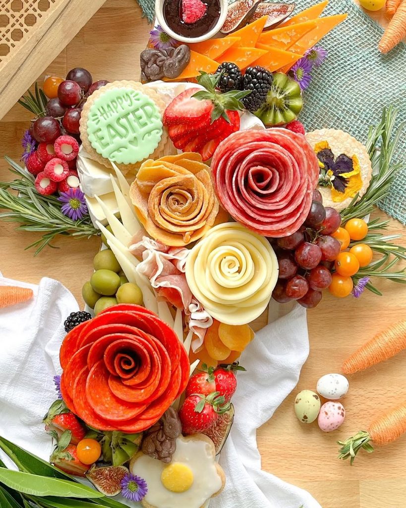 Spring-themed Easter charcuterie board featuring decorated Happy Easter cookie, salami roses, cheese roses, fresh berries, grapes, cheeses, chocolate dip, and edible flowers arranged with decorative carrots and speckled eggs
