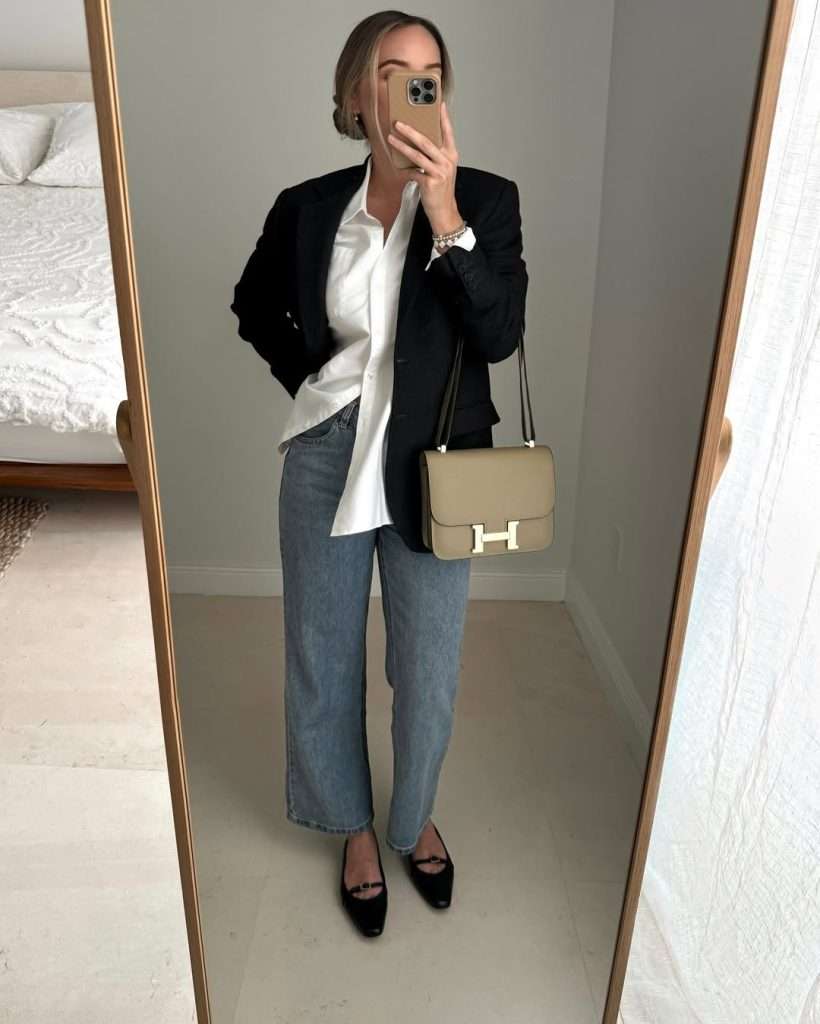 Business casual outfit with black blazer, white oversized button-down shirt, blue straight-leg jeans, black flats and beige designer shoulder 